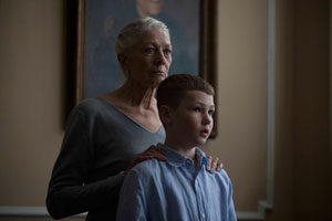 a mid shot with dark lighting shows an older woman standing with her hands on the shoulders of a young boy. Both ear serious expressions and are looking out to the front right of the shot.