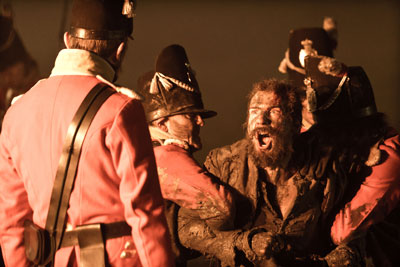 Mid shot of a muddy and raggedly dressed man struggling and shouting. Soldiers in red are restraining him