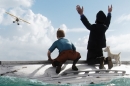 An animated boy and dog sit on a capsized boat whilst the man beside him tries to flag down a plane