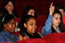 A row of girls sitting in a cinema, one with their hand in the air.