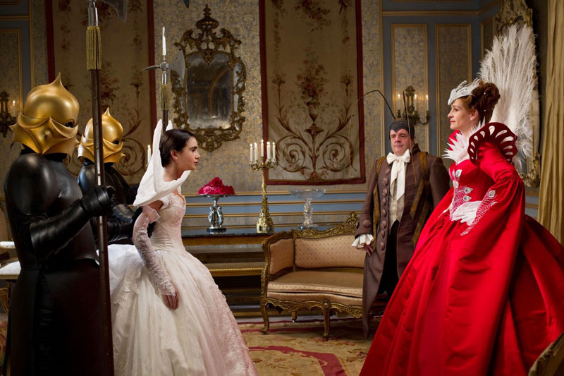 Five people stand in a smartly decorated room. There are two guards wearing shiny gold helmets that cover their faces. They are stood either side of a young woman dressed in a white gown. Opposite them is a man in period dress and a fancy dress 'antennae' hat. Next to him is a woman with her hands on her hips, wearing a scarlet dress with a white peacock tail attached to the back.