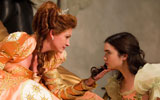 A woman wearing a tiara and a peach and gold gown is sitting. She is cupping the chin of a young woman who is kneeling down, looking up at her