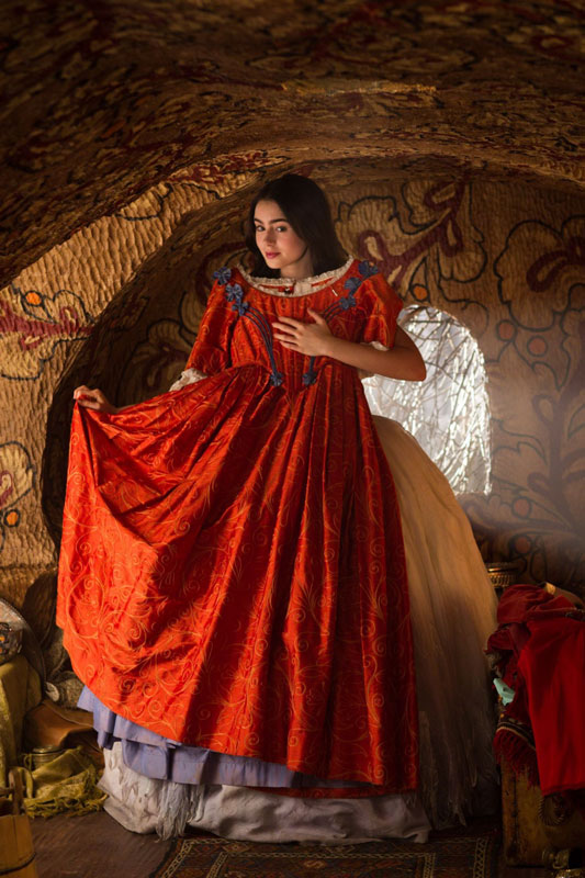 A young woman holds up a red gown against herself. She is standing in a cave-like dwelling with painted flowers decorating the walls.