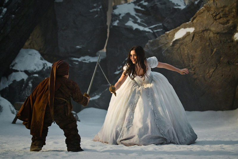 A snow scene. A young woman dressed in a white gown is having a sword fight with a dwarf.