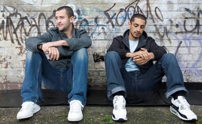 2 adolescent males sat with their backs against a wall, which has grafitti tags on it. Their appearence is that of tpical london youths, crewcut hair, jeans, white trainers and hooded tops.