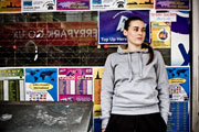 Mid shot of a young woman dressed in a grey hoodie, leaning against an empty shop front that is covered in posters