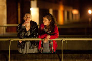 Mid long shot at night, showing a younger girl and a young woman leaning on the railings of a housing estate concrete walkway. The younger girls is looking at the older one who is smoking