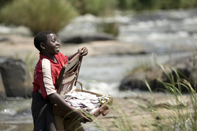 An African child stands next to a stream holding open a suitcase filled with banknotes.