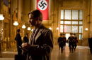 A man stands in a train station with a swastika on a banner behind him and SS officers in the background