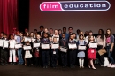 Lots of children of various ages stand on a stage with a man holding certificates and trophies.