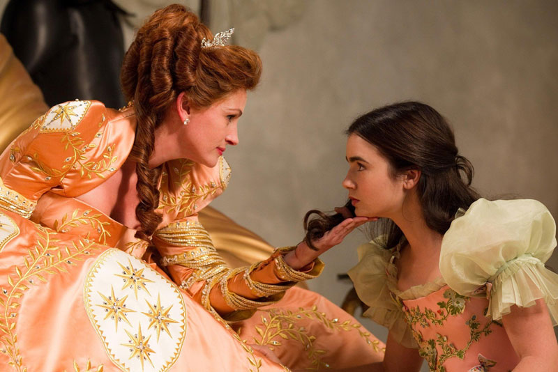 A woman wearing a tiara and a peach and gold gown is sitting. She is cupping the chin of a young woman who is kneeling down, looking up at her