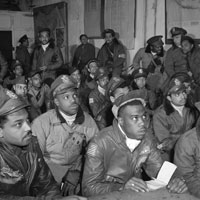 Large group of African American Tuskegee airmen attending a briefing, all wearing flight jackets with the US flag on their sleeves