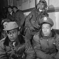 African American Tuskegee airmen attending a briefing; some are leaning against a wall; two in the foreground are looking to the right of the camera