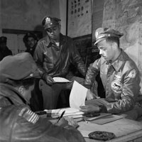 Two African American airmen sit either side of a desk, one is checking papers, a third watches from the side, sitting perched on a table