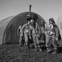 Several African American airmen in full flight suits leave the parachute room which looks like a large tent pitched on muddy terrain