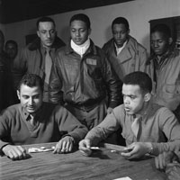 Two Tuskegee airmen playing cards at a table; several others in flight jackets stand behind watching the game