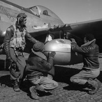 Two Tuskegee airmen wearing flight jackets crouching down attending to the wing of a plane. Another airman in full flight suit stands beside them