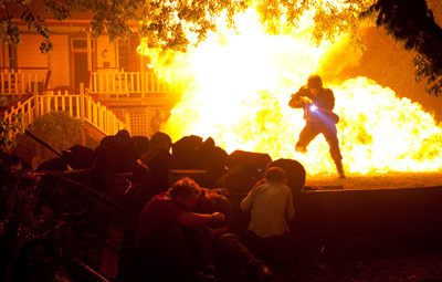 Three teenagers – two girls and a boy – crouch behind a log pile in the foreground of this long shot. At the rear of the shot, an armed uniformed soldier stands in the centre of a fiery blast.
